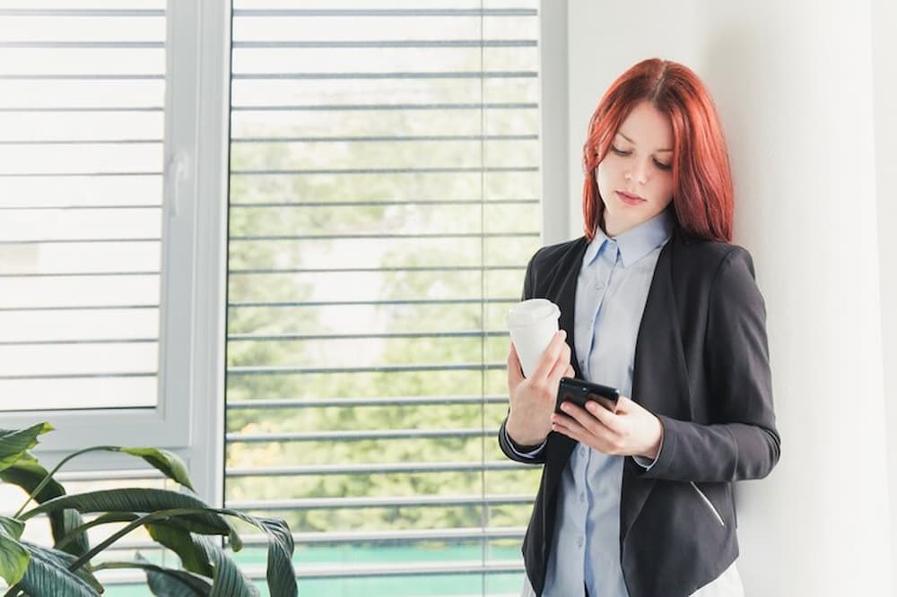 mujer viendo el móvil junto a ventana con persianas